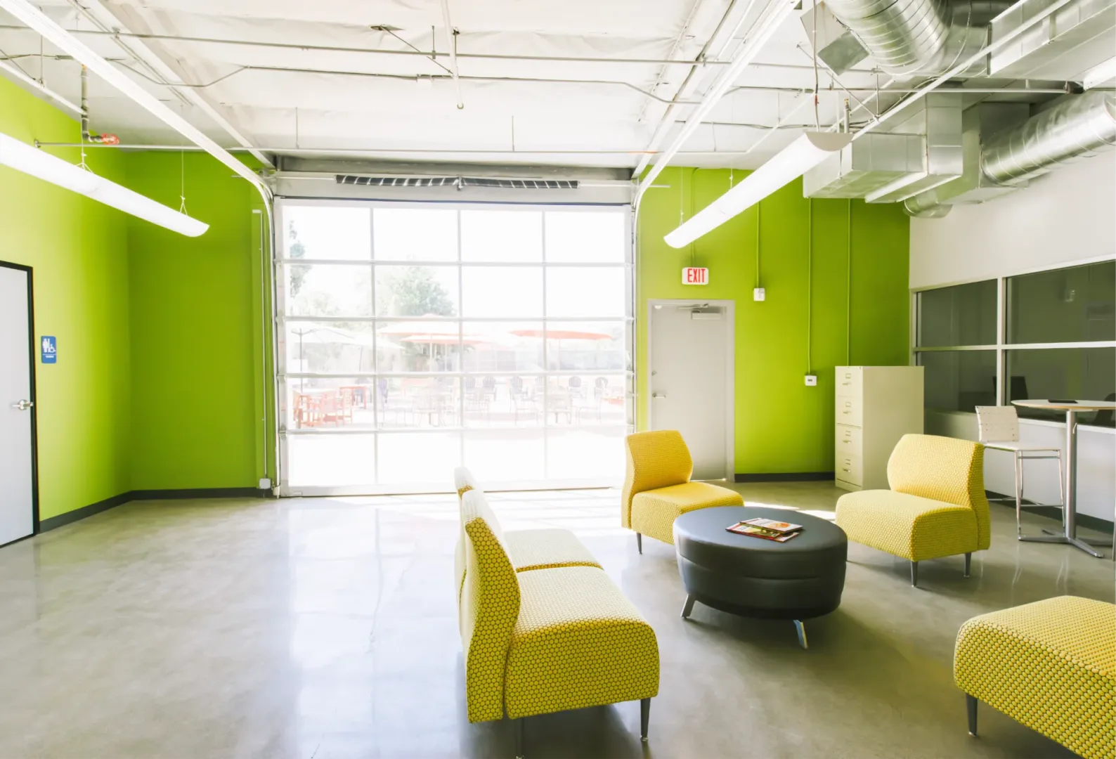 Colorful youth lounge with yellow chairs, lime green walls, and a garage-style window overlooking the outdoor patio at the Boys & Girls Club, designed by Visioneering Studios.