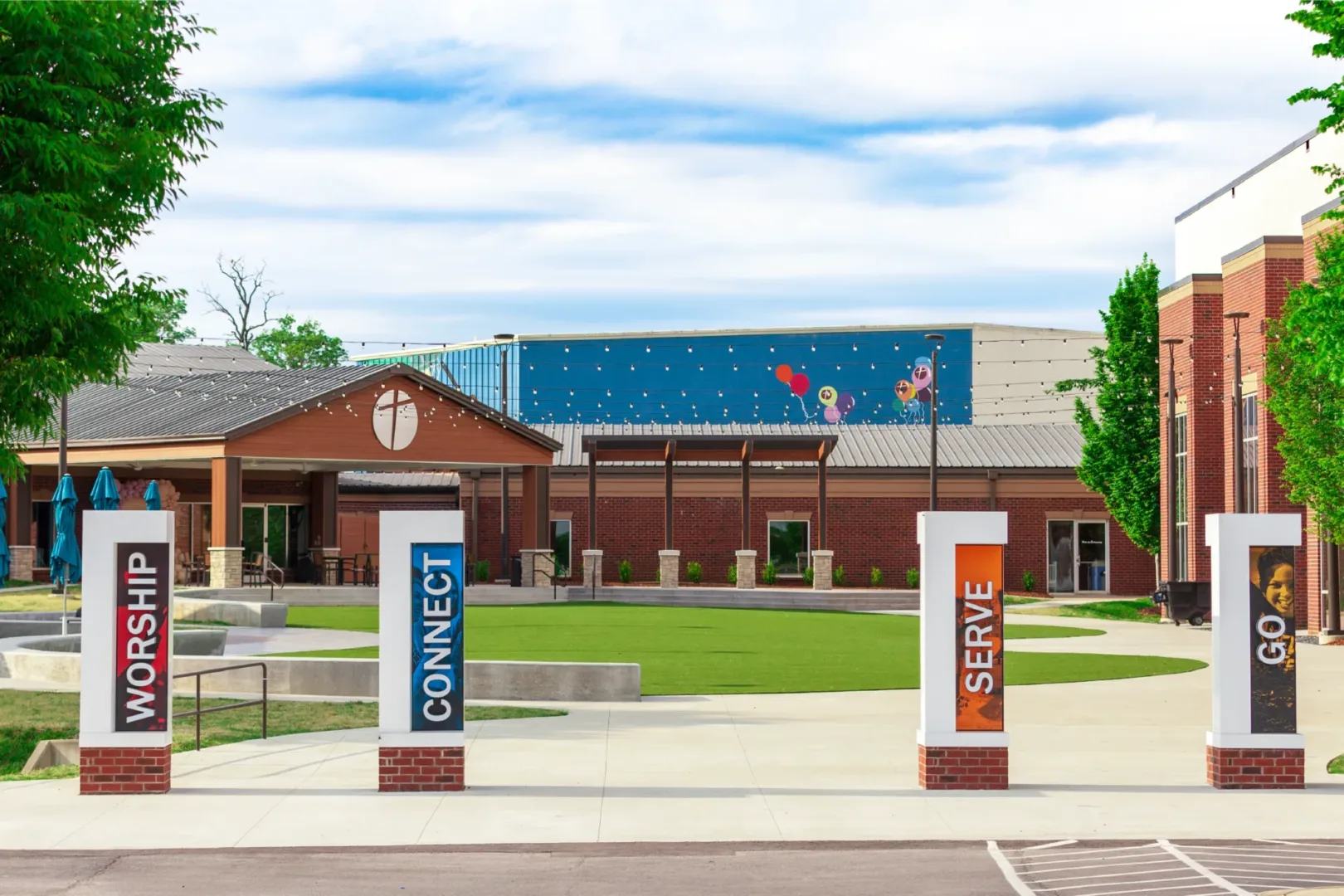 Exterior shot of New Vision Baptist Church’s Town Square, an open gathering area featuring pillars with Worship, Connect, Serve, Go messages near the Welcome 1 portal. church architect - visioneering studios