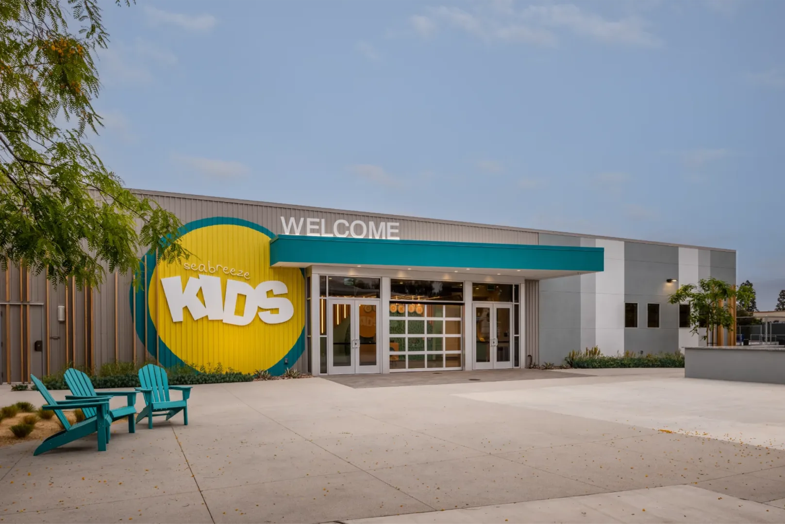 Exterior of Seabreeze Church’s secure kids building by church architect Visioneering Studios, featuring “Seabreeze Kids” signage, beach-style awning, blue lounge chairs, and visible check-in space through doors and garage-style window