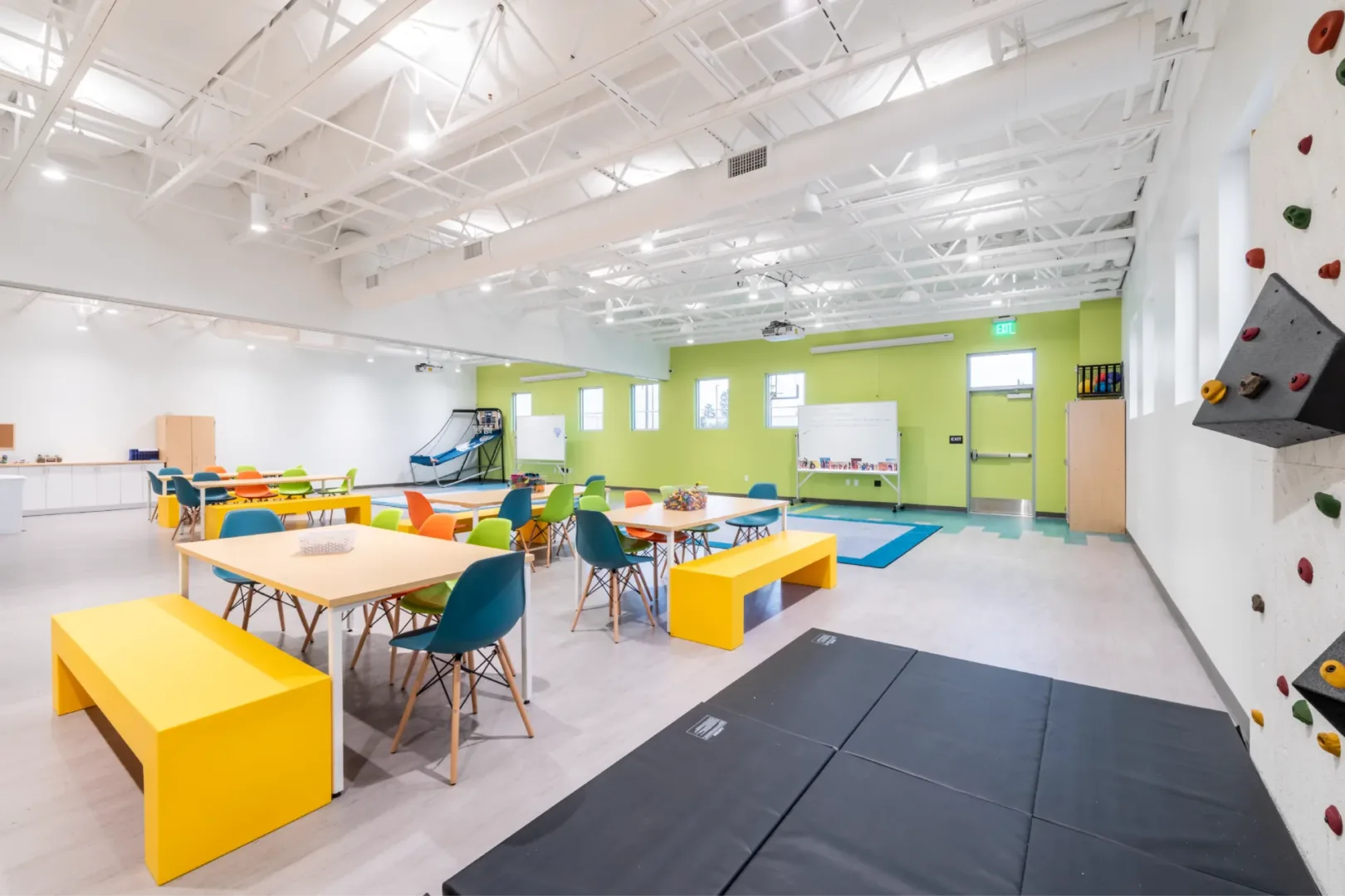 Large kids’ classroom at Seabreeze Church designed by church architect Visioneering Studios, featuring tables for group activities, an indoor basketball shooting game, a climbing rock wall, and a whiteboard area for reading or teaching time.
