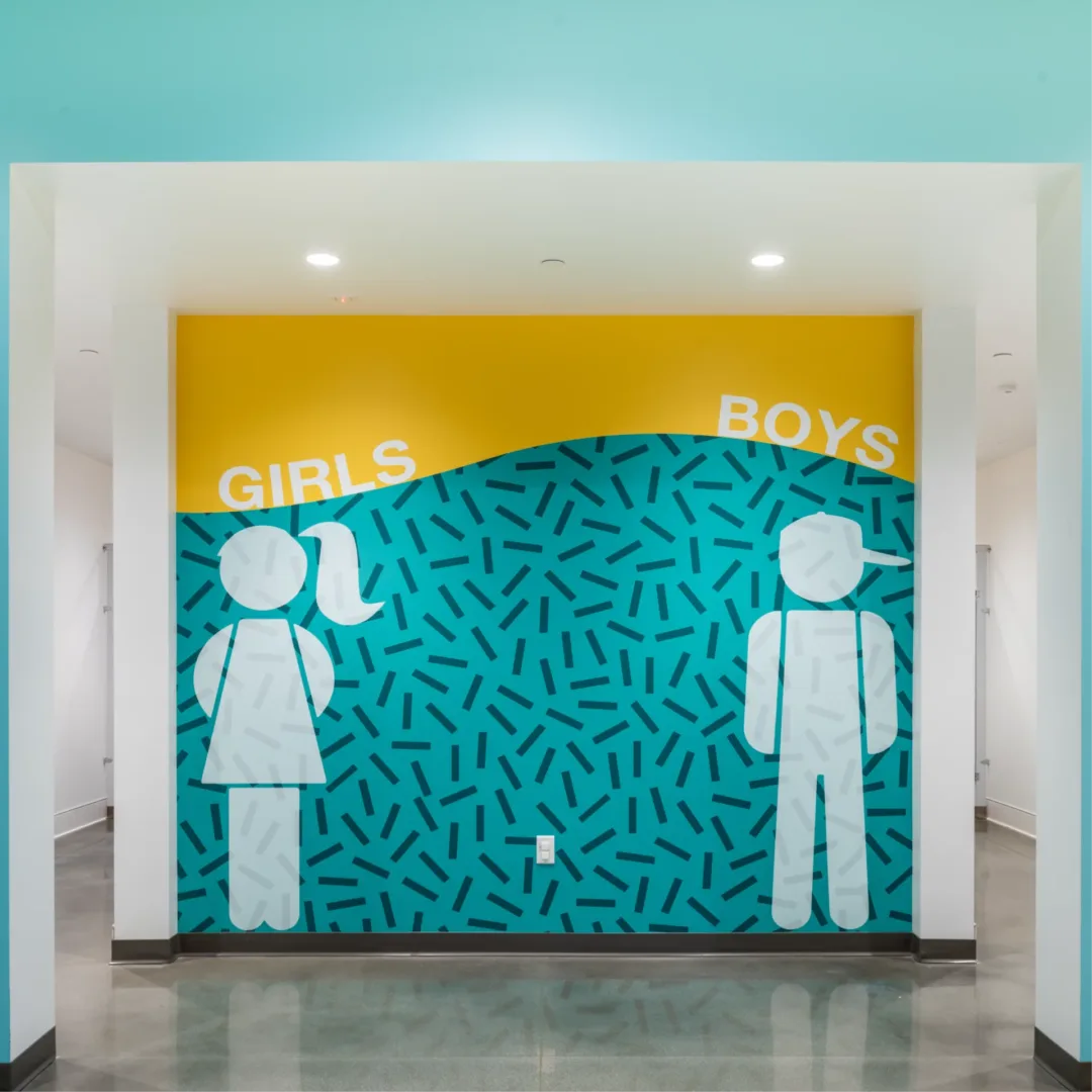 Interior wall graphic design at Seabreeze Church by Visioneering Studios, located outside the boys’ and girls’ restrooms. The design matches the kids’ check-in space with the same vibrant colors, featuring a playful large graphic of a boy with a sideways hat and a girl illustration to indicate restroom gender.