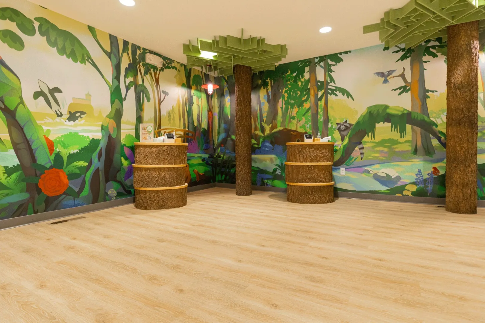 A vibrant, nature-inspired kids check-in lobby with immersive forest murals, faux-tree columns, custom log-style check-in stations, and playful environmental design by Visioneering Studios.