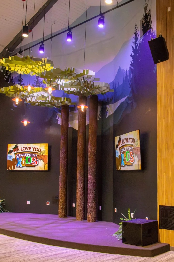 A themed kids large-group environment with faux tree trunks, layered canopy elements, a mountain mural backdrop, and Gracepoint Kids branding designed by Visioneering Studios.