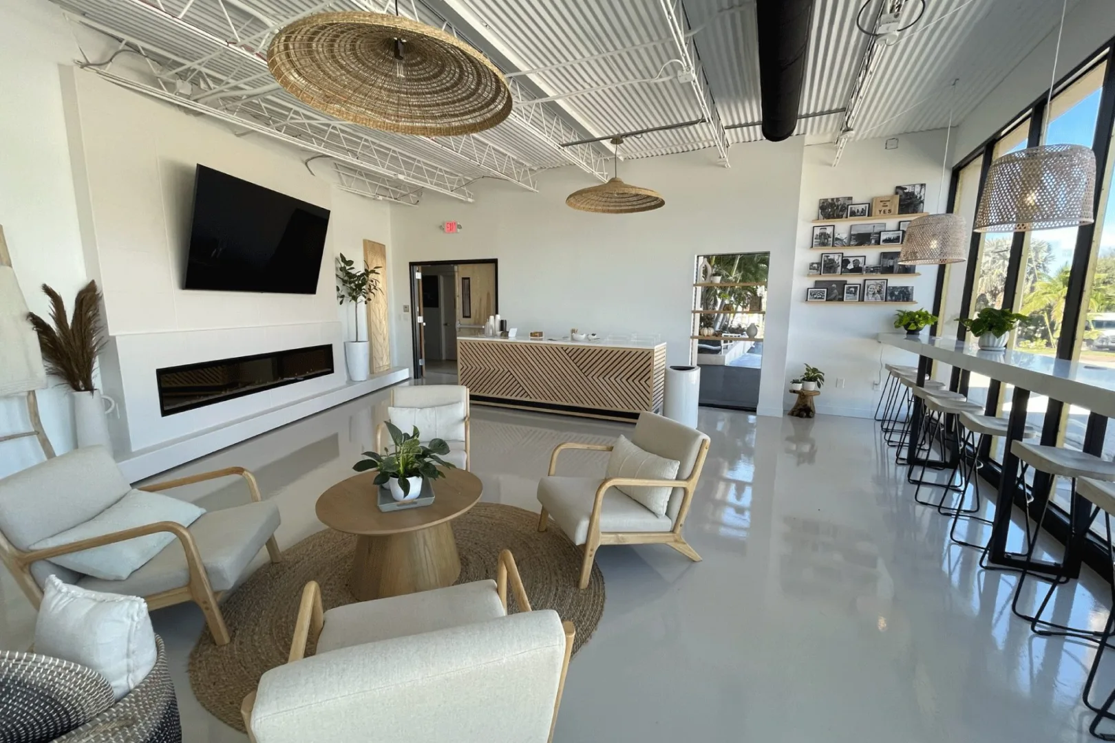 A bright, modern lobby featuring a café counter with wood chevron details, lounge seating, woven decor, and a display wall of framed photos.