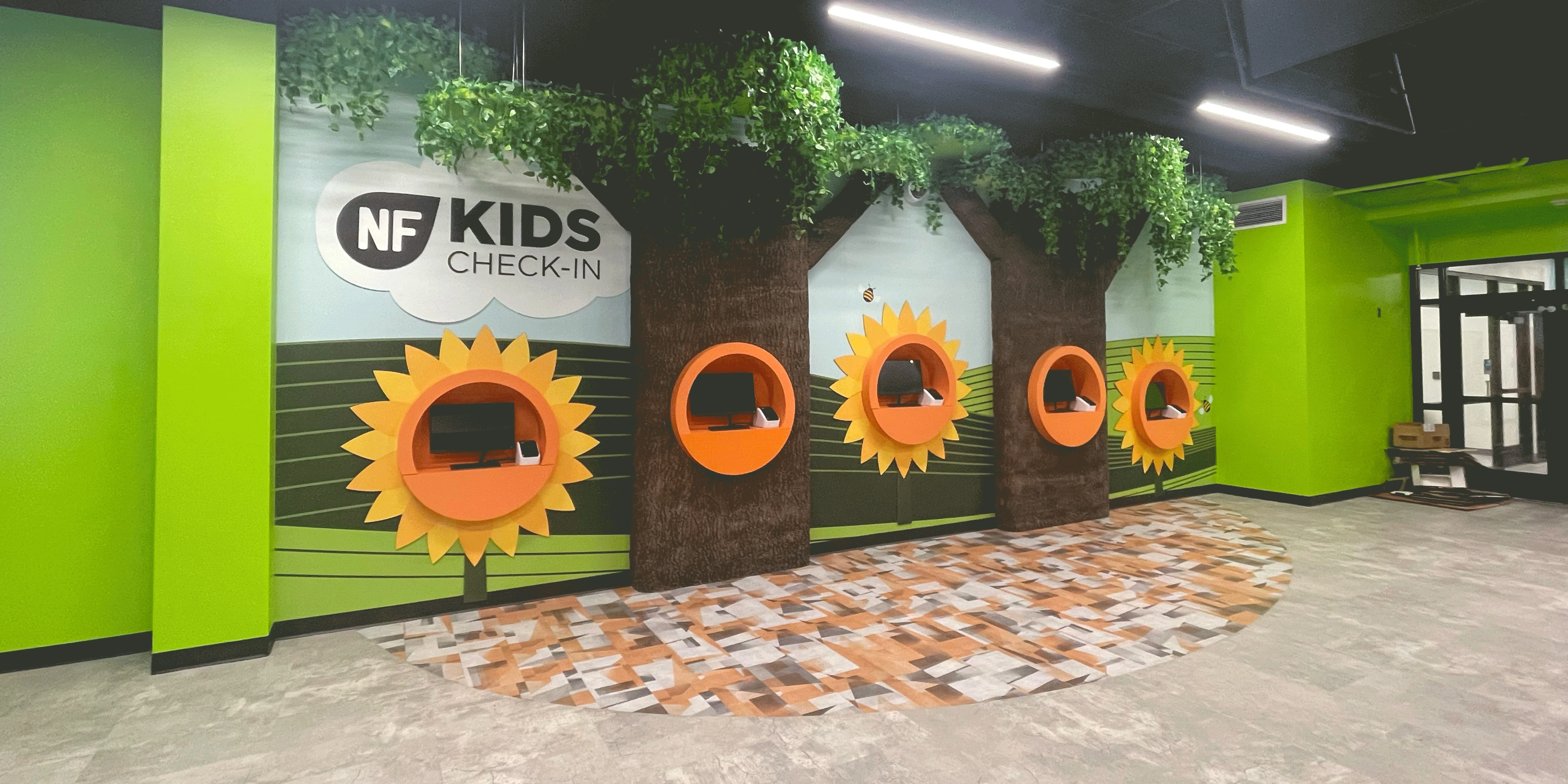 Completed northfield church kids ministry check-in area with custom tree-themed millwork, colorful environmental graphics, integrated technology kiosks, and wayfinding designed by church designer Visioneering Studios.
